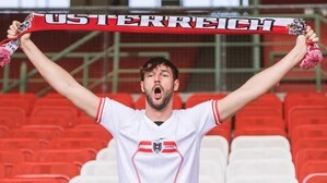 Ein Mann sitzt in einem Fußballstadion und hält einen Österreich-Schal in die Höhe.