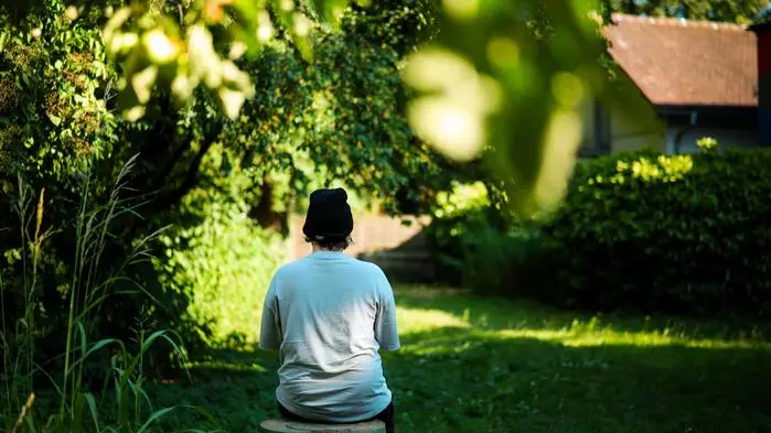 Ein junger Mann mit weißen T-Shirt und Haube sitzt auf einer Bank in der Wiese; fotografiert von hinten, um die Anonymität zu wahren
