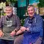 Zwei Männer sitzen vor Blumen, halten sich die Hände und lachen in die Kamera
