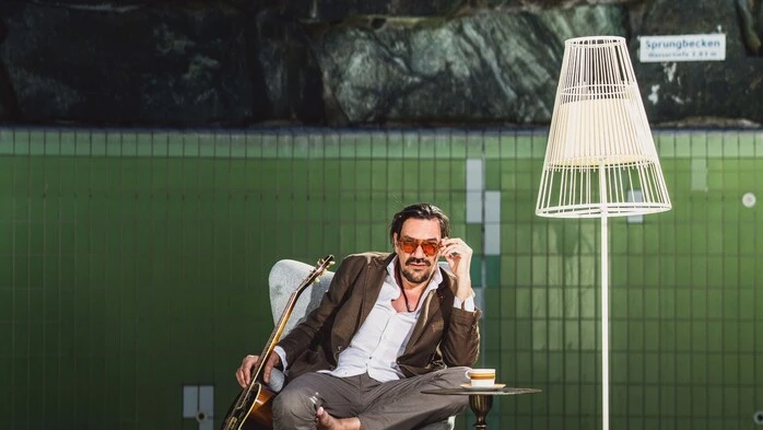 Ein Mann mit einer Gitarre sitzt auf einem Stuhl in einem leeren Schwimmbad.