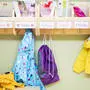 Sujetbild einer Garderobe in einem Kindergarten; es hängen Regenjacken und Turnbeutel, Namen wie Clara, Frieda und Paul stehen auf bunten Schildern