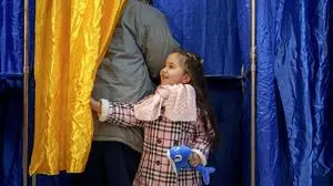 Ana Caroline, 3 and a half years-old, holds a toy while pulling the curtain of a voting booth as an adult votes in the country's parliamentary elections, in Bucharest, Romania, Sunday, Dec. 1, 2024. (AP Photo/Andreea Alexandru)