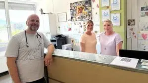 Ein Mann mit Glatze lehnt an einer Empfangstheke in einer Arztpraxis. Zwei blonde Frauen stehen dahinter.