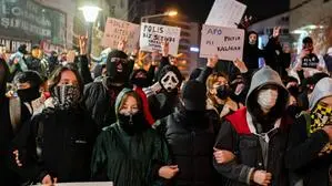 Das Bild zeigt eine Demonstration mit einer Gruppe von Menschen, viele davon vermummt und mit Masken, die in einer städtischen Umgebung protestieren. Mehrere Personen halten Schilder mit politischen Botschaften in türkischer Sprache hoch. Die Protestierenden tragen warme Kleidung und es findet offensichtlich bei Nacht statt. Die Stimmung wirkt angespannt, und einige Demonstranten zeigen Symbole oder Gesten mit ihren Händen. Die Inhalte der Schilder deuten auf politische Kritik und Forderungen hin. | Viele der sieben Millionen Studierenden in der Türkei sind wütend