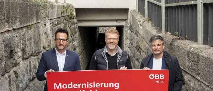 Drei Männer halten ein rotes Schild in die Kamera. Dort ist in großen Buchstaben „Modernisierung Knittelfeld“ zu lesen