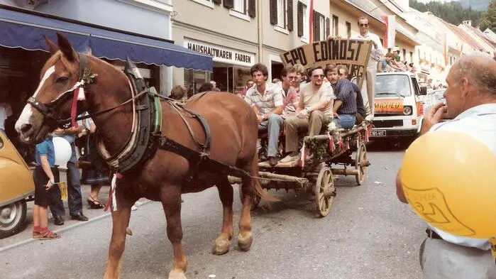 Zu sehen ist ein Umzug – Männer sitzen auf einem Wagen, der von einem Pferd gezogen wird. 