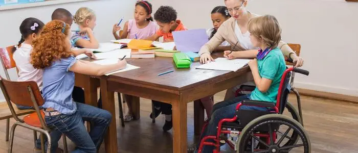 Bild zeigt eine Gruppe von Volksschulkindern an einem Tisch mit einer Lehrerin, ein Mädchen sitzt im Rollstuhl