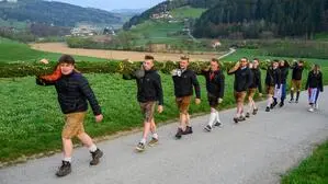 Junge Männer in Tracht gekleidet spazieren auf der Straße zwischen Wiesen mit einem Palmbuschen auf den Schultern