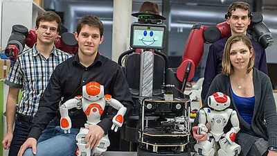 Baxter, der Roboter mit Hut und Schlips, umschlingt seine Erzeuger Markus Plass, Stefan Imlauer, Julia Nitsch und Emilian Jungwirth (v.l.). Er hat ein Lächeln im Gesicht und freut sich auf seinen ersten Auftritt am Freitag im Congress.