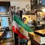 Ein Mann steht im Innenraum einer Bar und spannt vor seinem Körper eine große Flagge des Iran auf