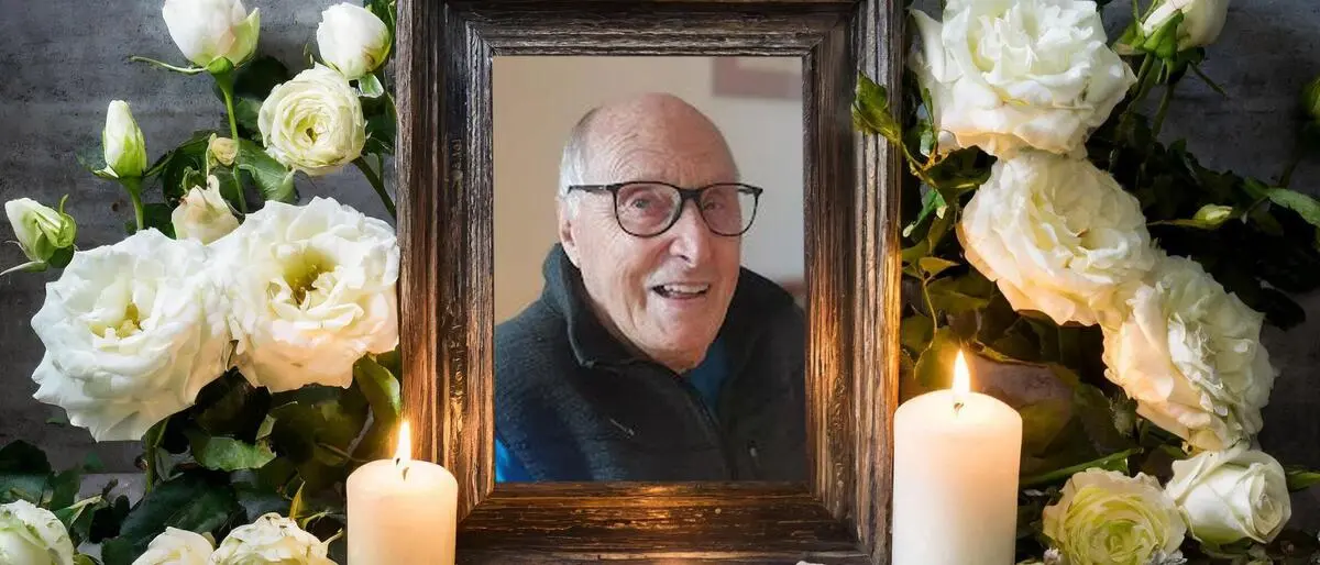 Ein gerahmtes Bild von einem alten Mann steht auf einem Tisch. Der Rahmen ist geschmückt mit weißen Rosen, Kerzen brennen im Vordergrund