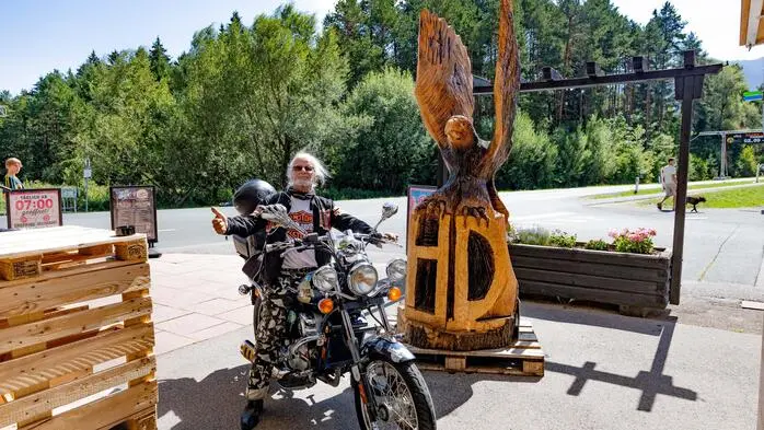 Ein Mann mit weißen Haaren und „Harley-Outfit“ posiert auf seinem Moped, dahinter ein Straße und ein Wäldchen
