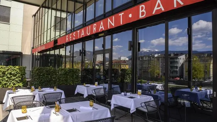 Terrasse eines Restaurants, mehrere Tisch mit weißer Tischdecke, dahinter eine Glasfront mit der Aufschrift „Restuarant, Bar“