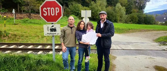 Eine dreiköpfige Familie steht an einem Bahnübergang. Daneben steht ein Mann mit einer Baskenmütze auf