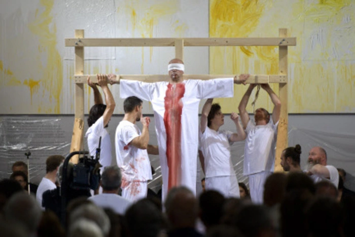 Langsam wird ein auf einem Holzgerüst mit BändernGekreuzigter im weißen Hemd in den oberen Bereich des Raums getragen. Auf dem Boden zertreten die Akteure Paradeiser und Trauben zu Sugo.