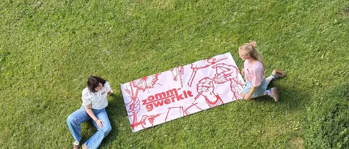 Ein großes Plakat liegt auf einer Wiese, zwei Frauen sitzen links und rechts davon