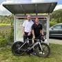 Ein Mitte 50-jähriger Mann im Hemd steht neben einem 39-jährigen Mann im Fahrradanzug. Beide halten sie ein Rennrad, das vor ihnen steht. Im Hintergrund sieht man die Landschaft von Himmelberg an einem sonnigen Tag