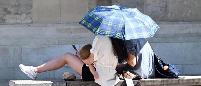Eine Familie sitzt auf einer betonierten Bank in einer Stadt unter einem Schirm, auf den die Sonne strahlt