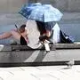 Eine Familie sitzt auf einer betonierten Bank in einer Stadt unter einem Schirm, auf den die Sonne strahlt