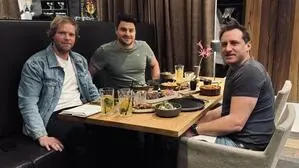 Drei Männer sitzen in einem Restaurant am Tisch, der gedeckt ist und lächeln in die Kamera