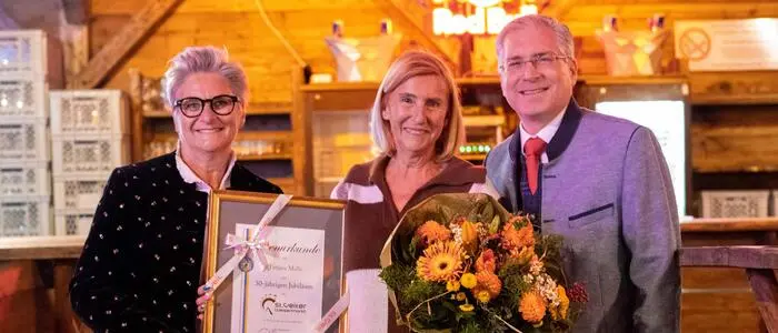 Zwei Frauen und ein Mann posieren für ein Foto. Die Frau in der Mitte hält einen Blumenstrauß in der Hand, die Frau links eine Urkunde.