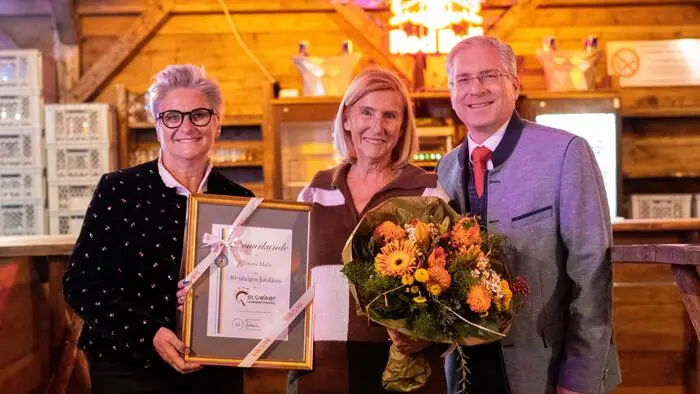 Zwei Frauen und ein Mann posieren für ein Foto. Die Frau in der Mitte hält einen Blumenstrauß in der Hand, die Frau links eine Urkunde.