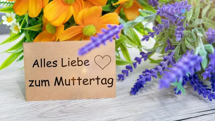 Eine graue Karte steht vor zwei Arten von Blumen, darauf steht „Alles Liebe zum Muttertag“