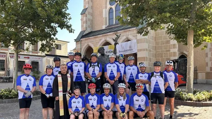 Eine Gruppe von Radfahrenden in Trikots vor einer Kirche und einer Statue des Heiligen Franziskus