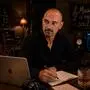 Ein Mann mit Glatze und Bart sitzt in einem dunklen Raum. Vor ihm ist ein Laptop. In seiner Hand ist ein Stift und neben ihm ist ein Stück Papier.