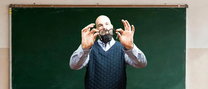 Ein Mann steht vor einer Tafel in einem Klassenraum und betrachtet eine zersplitterte Brille in seinen Händen