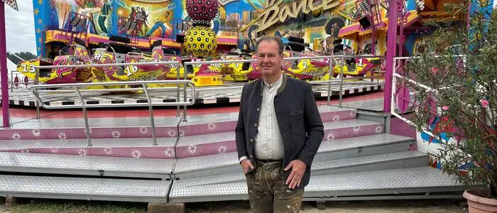 Andreas Lackner steht vor dem Fahrgeschäft Break Dance am St. Veiter Wiesenmarkt. Er trägt eine knielange Lederhose und einen Trachtenjanker