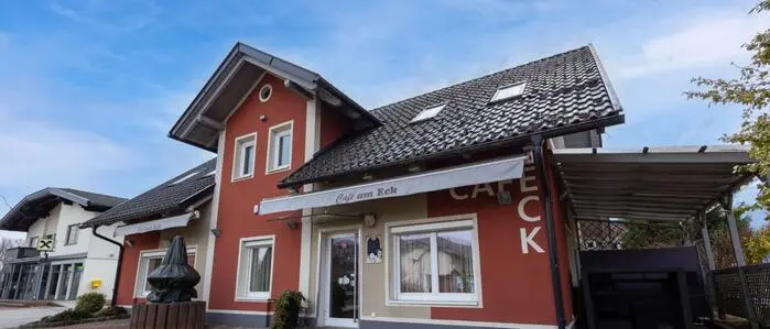 Seitlicher Blick auf ein rotes Haus, an der Fassade die Aufschrift „Cafè Eck! daneben ist ein leeres Carport