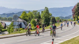 Viele Radfahrer fahren auf einer Straße, im Hintergrund sind Berge und ein See