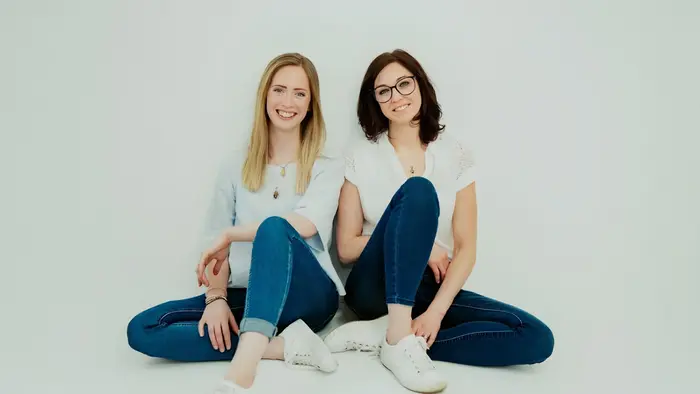 Zwei Frauen sitzen am Boden nebeneinander und lachen in die Kamera