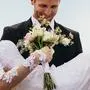Happy groom carrying beautiful bride with flowers in hand. Newlywed couple with bouquet outdoors on their wedding day.