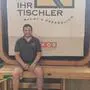 Ein Mann sitzt vor einem Schild „Ihr Tischler“