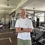 Ein blonder Mann in weißem Shirt steht in einem Fitnessstudio und lächelt in die Kamera