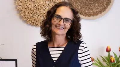 Eine Frau mit dunklen Locken, einer Brille und gestreiftem T-Shirt lacht in die Kamera