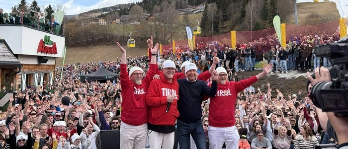 Vier Männer stehen auf einer Bühne. Im Hintergrund sind Tausende Fans zu sehen.