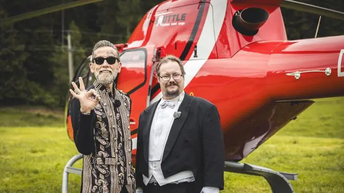 Ein Mann im extravaganten Outfit steht vor einem Helikopter. Neben ihm sein Butler in einem Frack. Der Mann signalisiert „OK“ in Richtung Kamera.