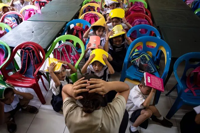 Die Kinder führen die Anweisungen der Instruktorin aus: Am Boden sitzend ducken sie sich zwischen Stühle, um die Chance, von umherfliegenden Gegenständen getroffen zu werden, zu minimieren.