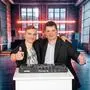 Zwei junge Männer vor einem DJ-Pult in einer Partylocation. Beide halten einen Daumen nach oben