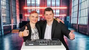 Zwei junge Männer vor einem DJ-Pult in einer Partylocation. Beide halten einen Daumen nach oben