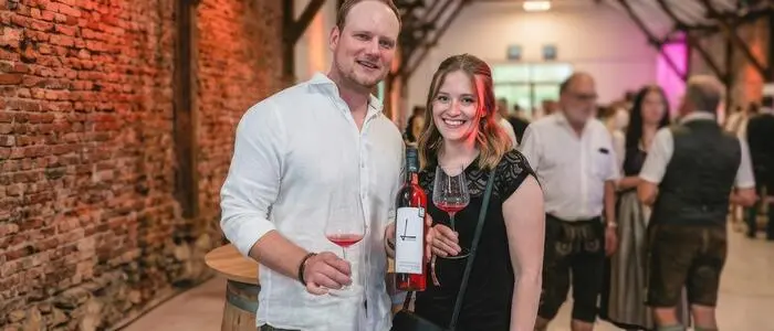 Ein Mann und eine Frau lachen in die Kamera. Der Mann hält eine Flasche Wein und ein Weinglas in seinen Händen.