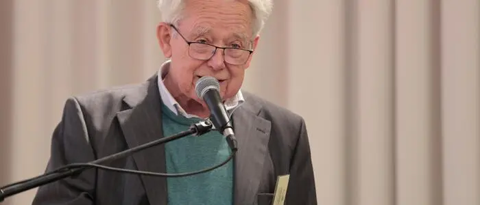 Man sieht einen grauhaarigen Mann an einem Pult mit Mikrofon sitzen