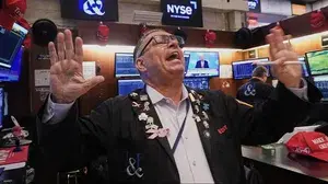 Ein Börsenhändler hebt gestikulierend die Hände, während hinter ihm Bildschirme mit Kursdaten und Nachrichten laufen. | Trump verzichtet auf Strafzölle gegen die EU, die Börsen freut es