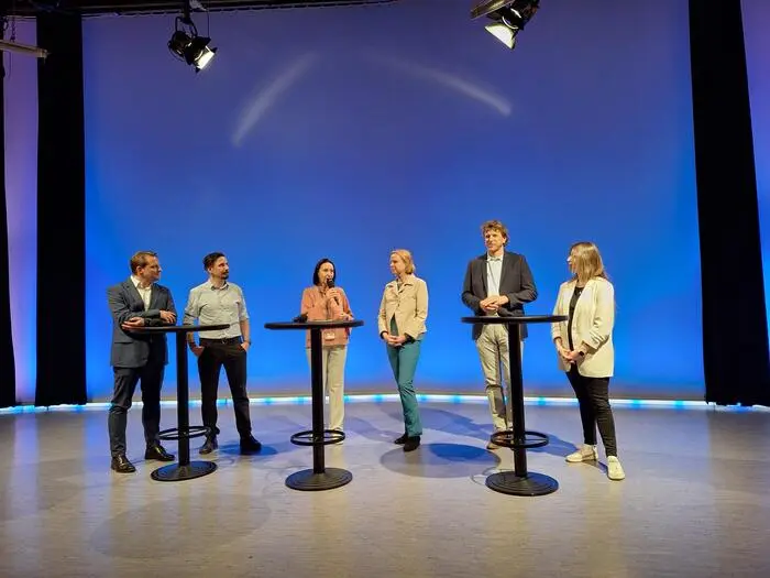 Sechs Personen stehen vor einem blauen Hintergrund, eine Frau spricht in ein Mikrofon