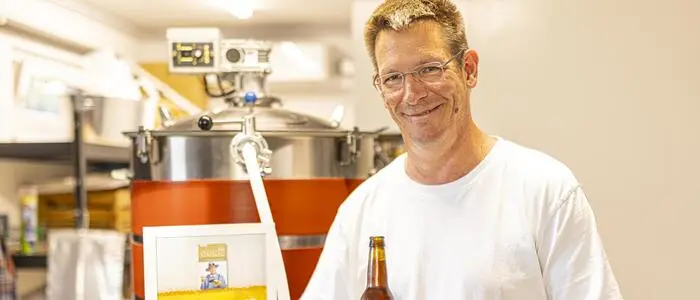 Gunther Kühnelt hält eine Urkunde und eine Bierflasche in der Hand und steht vor einem Braukessel