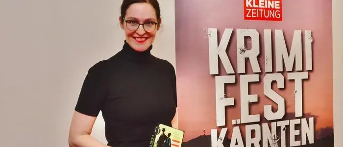Die Autorin steht mit einem Buch in der Hand vor einem Plakat | Alex Beer las im Falkensteiner in Tröpolach