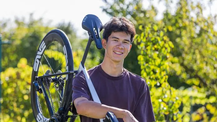 Ein junger Mann schultert ein Rennrad und lächelt in die Kamera, hinter ihm grüne Büsche